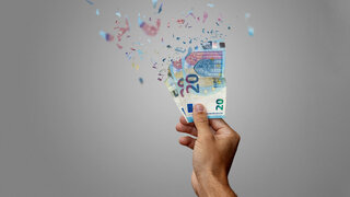 Eine Hand hält mehrere 20-Euro-Scheine, die sich in bunte Partikel auflösen, symbolisch für die Finanznot der Krankenkassen.