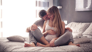 Ein Mann und eine schwangere Frau sitzen zusammen auf einem Bett, symbolisch für das Thema Sex in der Schwangerschaft.