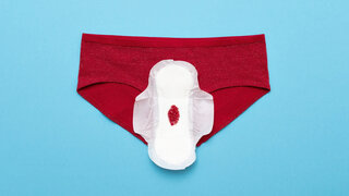 Ein roter Slip liegt auf einer blauen Fläche, darauf eine Binde mit einem kleinen Fleck Menstruationsblut.