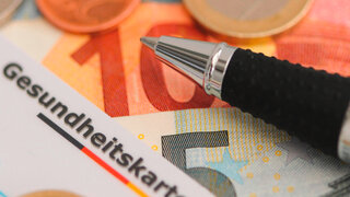 Eine Gesundheitskarte liegt auf Euro-Scheinen und Münzen, daneben ein Kugelschreiber, symbolisch für das Thema Einsparungen bei Krankenkassen.