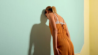 Eine Frau in einem orangefarbenen Kleid lehnt ihren Kopf gegen eine pastellgrüne Wand, während ihr Schatten deutlich auf die Wand fällt, symbolisch für psychisches Wohlbefinden.